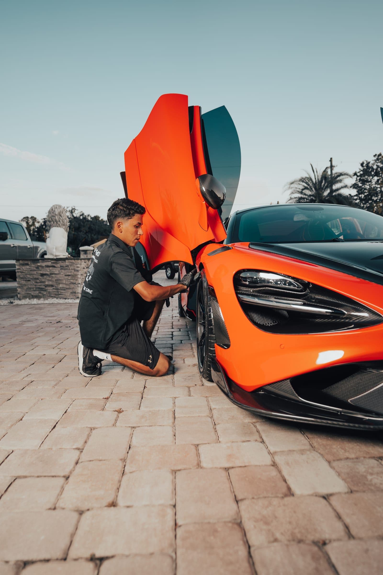 Technician kneeling to hand-detail the front fender and headlight of an orange McLaren with its dihedral door raised